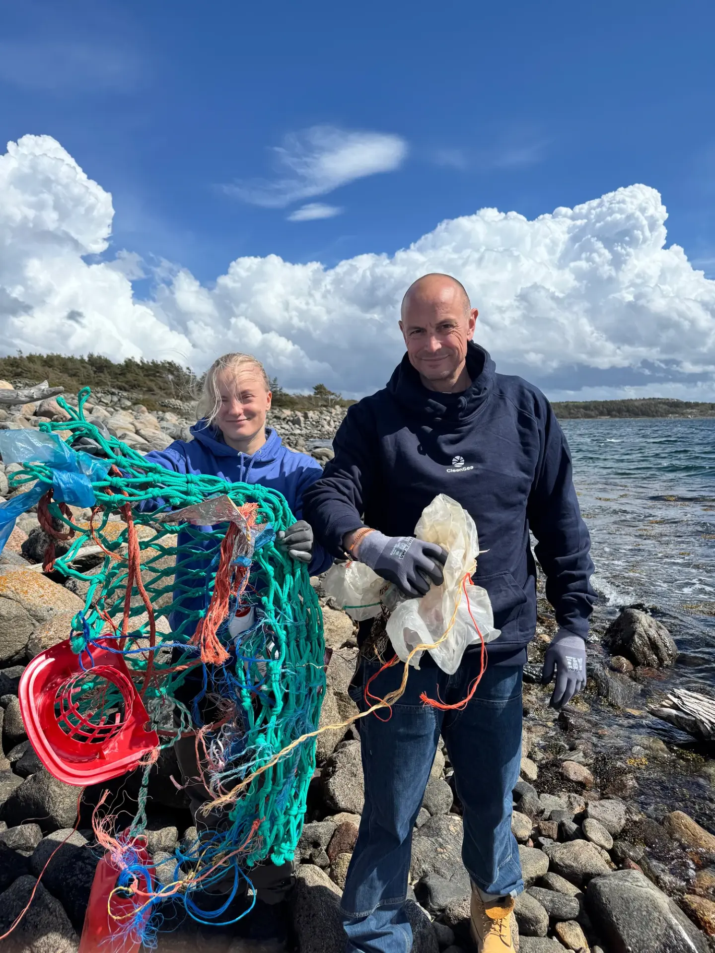 Två personer på en stenig strand håller upp en stor fångst av plastskräp och fiskenät
